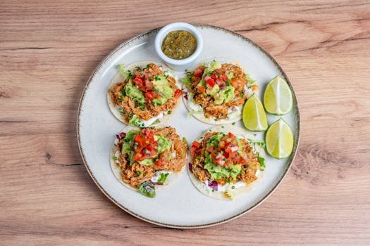 Four tacos topped with guacamole, diced tomatoes, and limes, served on a wooden surface.